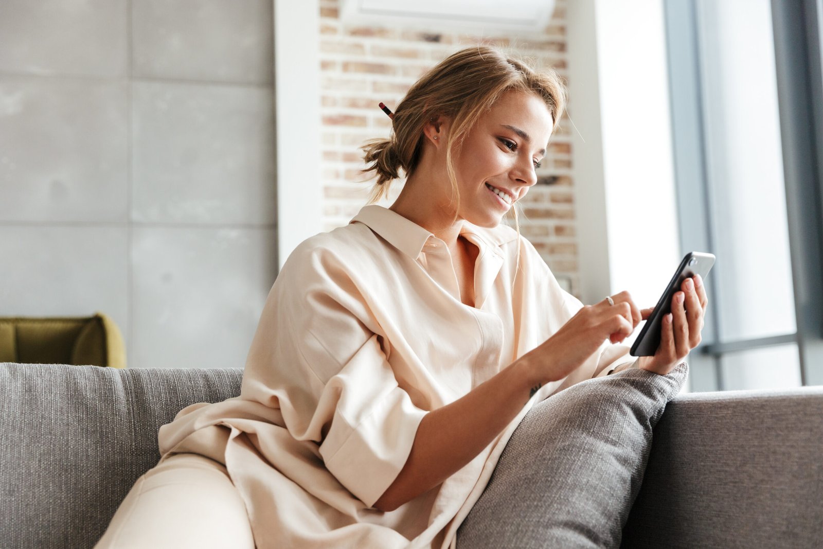 Image of happy woman smiling and using cellphone while sitting on sofa
